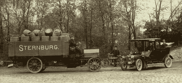Two old vehicles are standing on an avenue, one is a truck full of barrels, the other is a car.